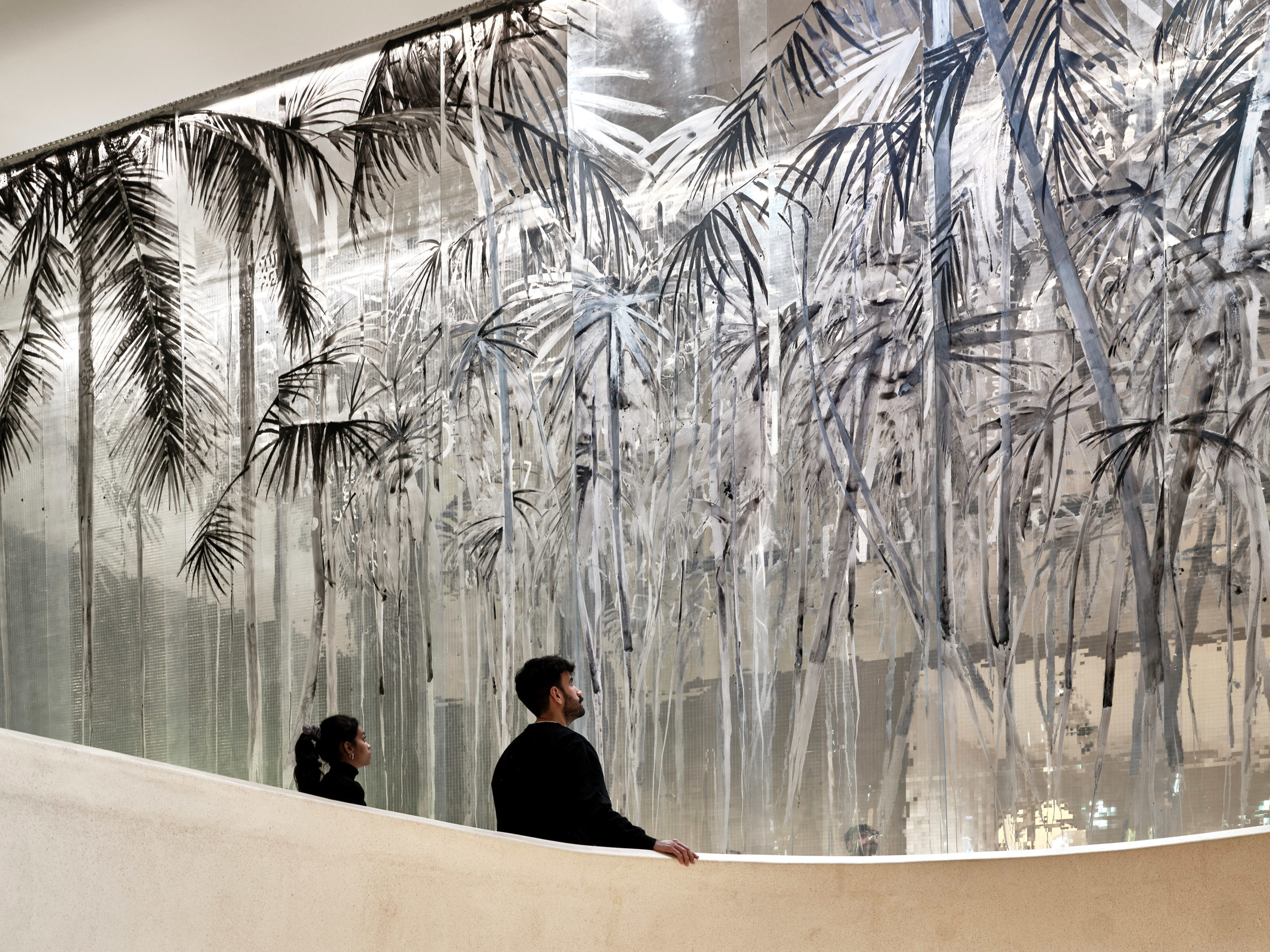 Ausstellungsansicht „Philipp Fürhofer. Phantominseln“ 
Zwei Menschen vor dem Vorhang der "Phantominseln": weiße bis Schwarze Palmsilhouetten die durch die Spiegelreflektion wie ein aschebdeckter Wald wirken.